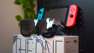 XREAL One AR glasses on top of a its packaging box featuring the words 'The One,' a Nintendo Switch 2 is in the background, out of focus.