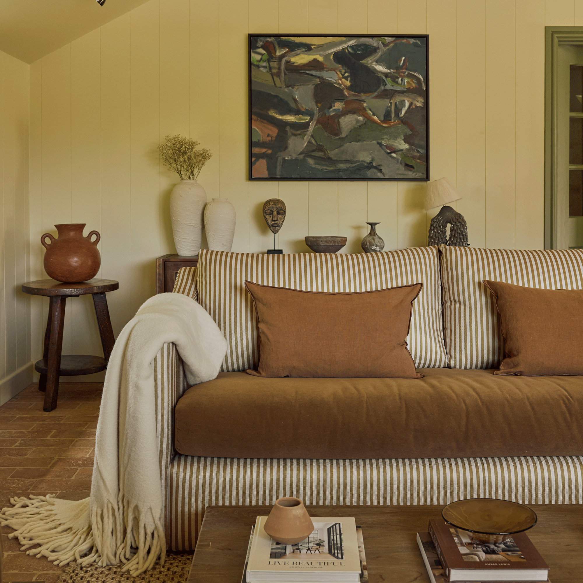 a yellow living room with a large sofa with mustard/brown cushions and striped upholstery, artwork on the wall and a coffee table with books
