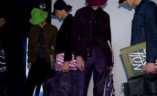 Five male models wearing looks from the Burberry Prorsum collection. One model is wearing a black coat. Another model is wearing black trousers and a denim piece with a green brown coloured jacket over the top. The third model is wearing a black jacket. The fourth model is wearing purple trousers and a black buttoned piece with a dark purple jacket over the top. And the fifth model is wearing a denim jacket. All models are wearing hats and holding notebooks with some holding briefcases