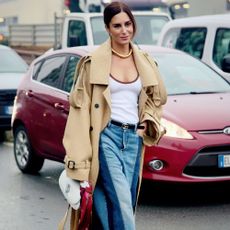 Woman wears white top, trench coat, and jeans while walking and posing on the street with cars behind her. She also carries a white handbag with a scarf tied around it. 