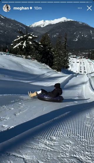 Meghan Markle wearing a black coat and hat riding a snow tube down a mountain