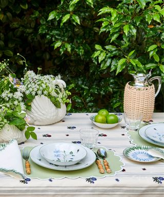 An outdoor dining table with blue patterned tablecloth with swan-shaped vases and a beige umbrella