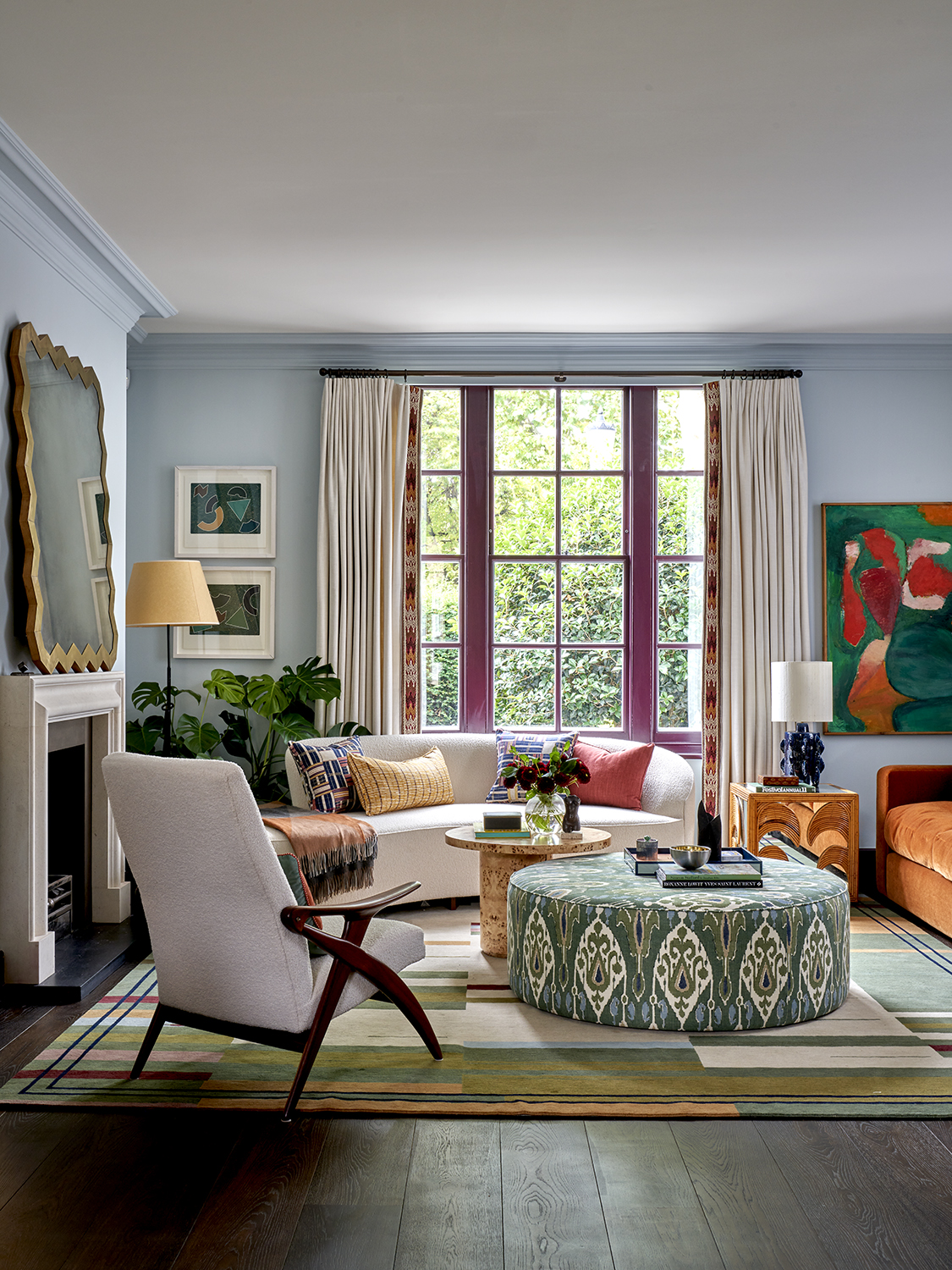 colorful living room with blue walls, white ceiling, dark floorboards, and white sofa with colorful throw pillows, a patterned upholstered round ottoman and side table, a white armchair with timber frame, a colorful rug, a fireplace with a mirror styled above and a window with fabric drapes