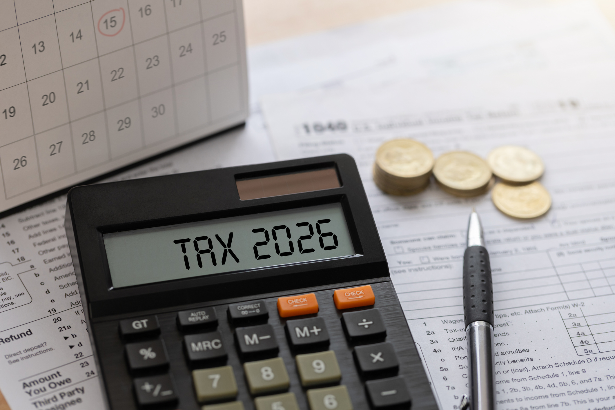 Annual tax planning concept with calculator displaying TAX 2026, stacks of coins, a calendar, and a pen on a wooden desk.
