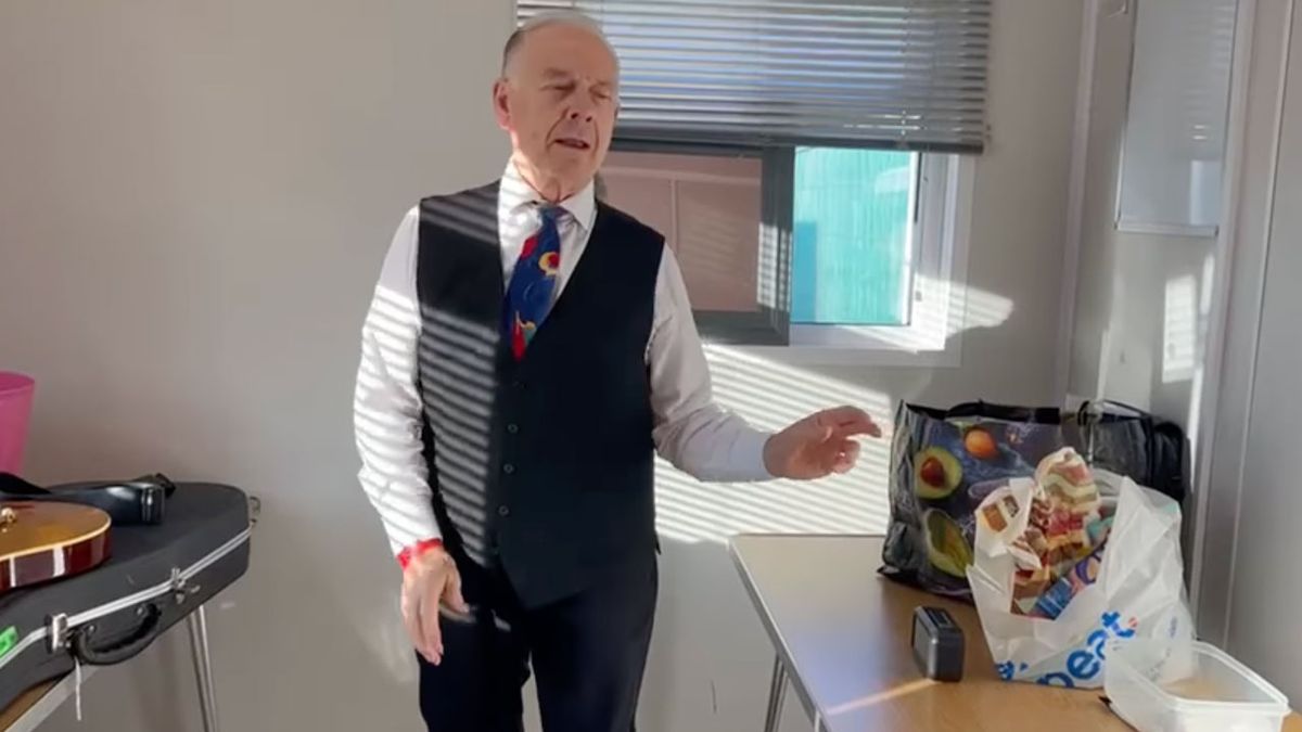 King Crimson&#039;s Robert Fripp dressed in a white shirt, colourful tie, dark waistcoat and trousers, with one arm outstretched mid-dance. He&#039;s photographed backstage in a small white dressing room with his guitar on a table to the left of the photo and a carrier bag of food to the right of the image