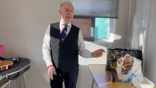 King Crimson's Robert Fripp dressed in a white shirt, colourful tie, dark waistcoat and trousers, with one arm outstretched mid-dance. He's photographed backstage in a small white dressing room with his guitar on a table to the left of the photo and a carrier bag of food to the right of the image