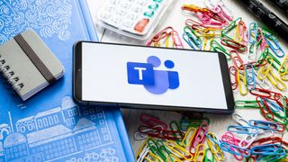 Microsoft Teams classic app logo pictured on a smartphone sitting on a desk with notepad and paper clips.