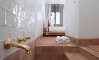 Interior view of a bathroom at Casa Modesta featuring white painted wall tiles, terracotta tiled flooring and steps, gold tap and handles, two rolled towels and soap on a dish. There is also a partial view of the bedroom