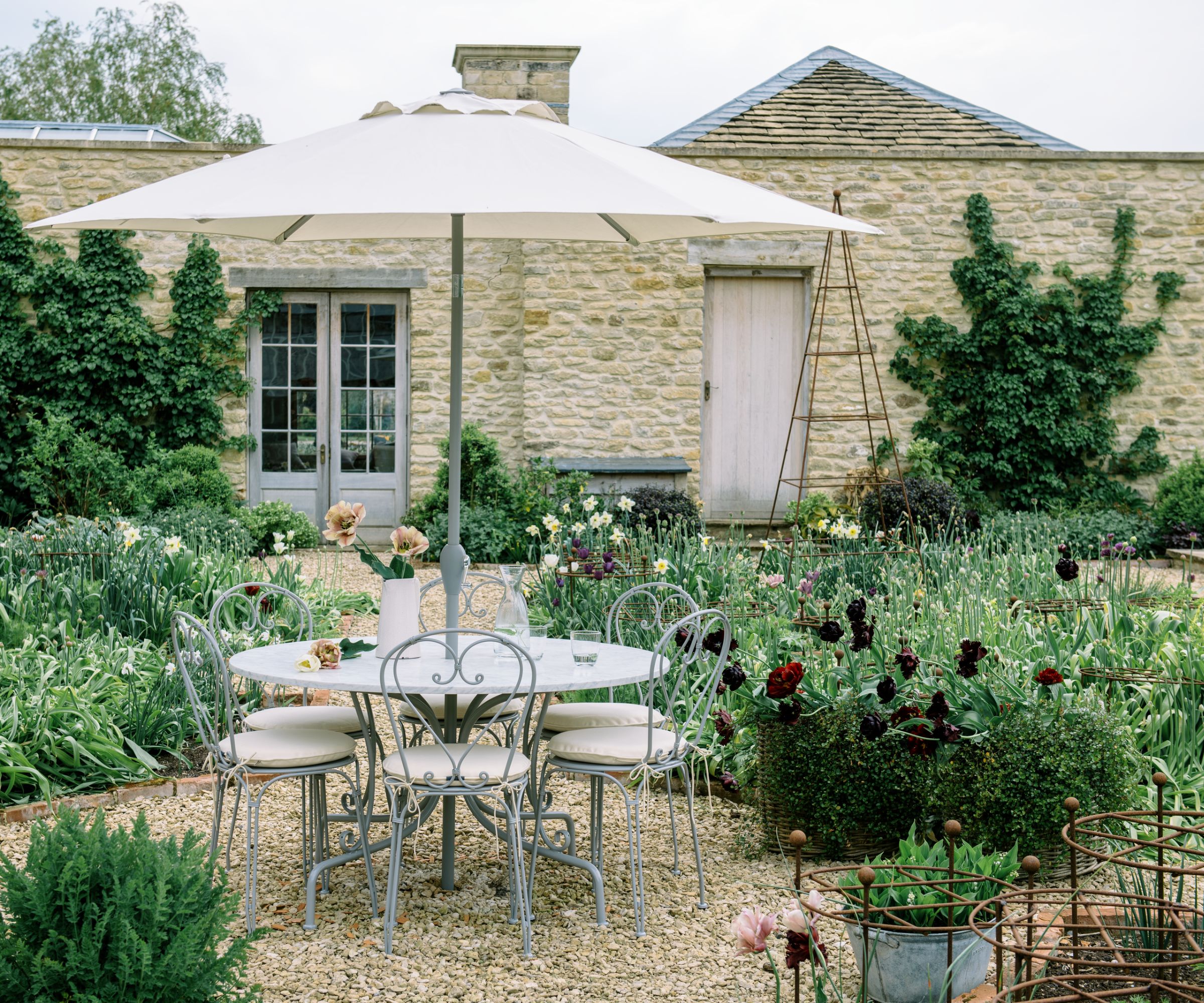 pale yellow stone walled garden with shingle stones dotted with plants and ornate wrought iron furniture with a patio umbrella