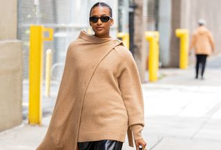 A woman wearing a tan scarf sweater, a leather skirt, and a black bucket bag.