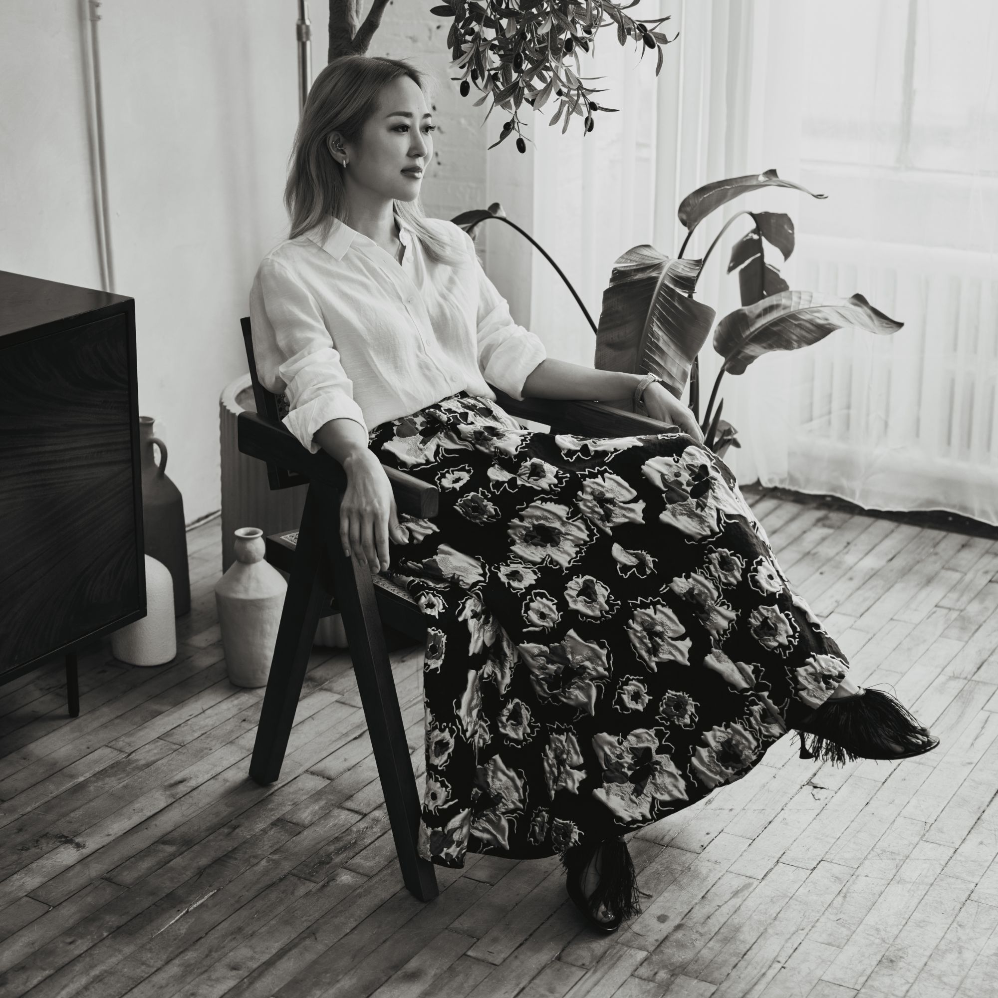 A black and white image of Maiko Shimazaki wearing a white skirt and black and white patterned skirt, sitting in a chair.