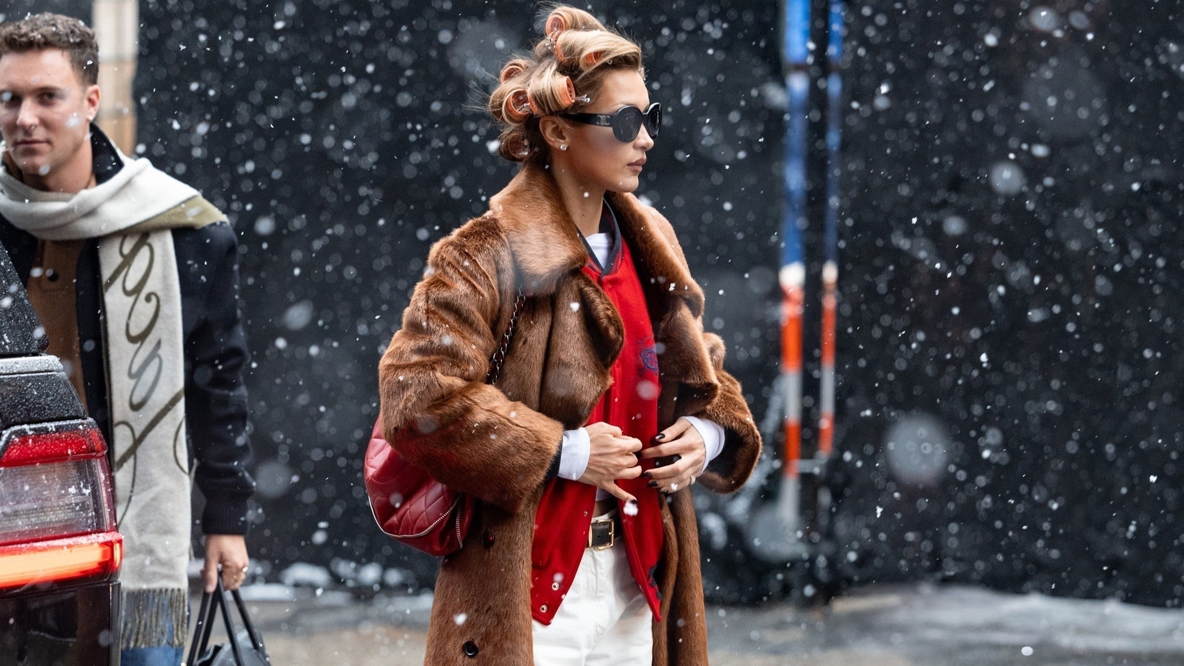 Bella Hadid wears white jeans with a red sweater, fur coat, and burgundy bag.
