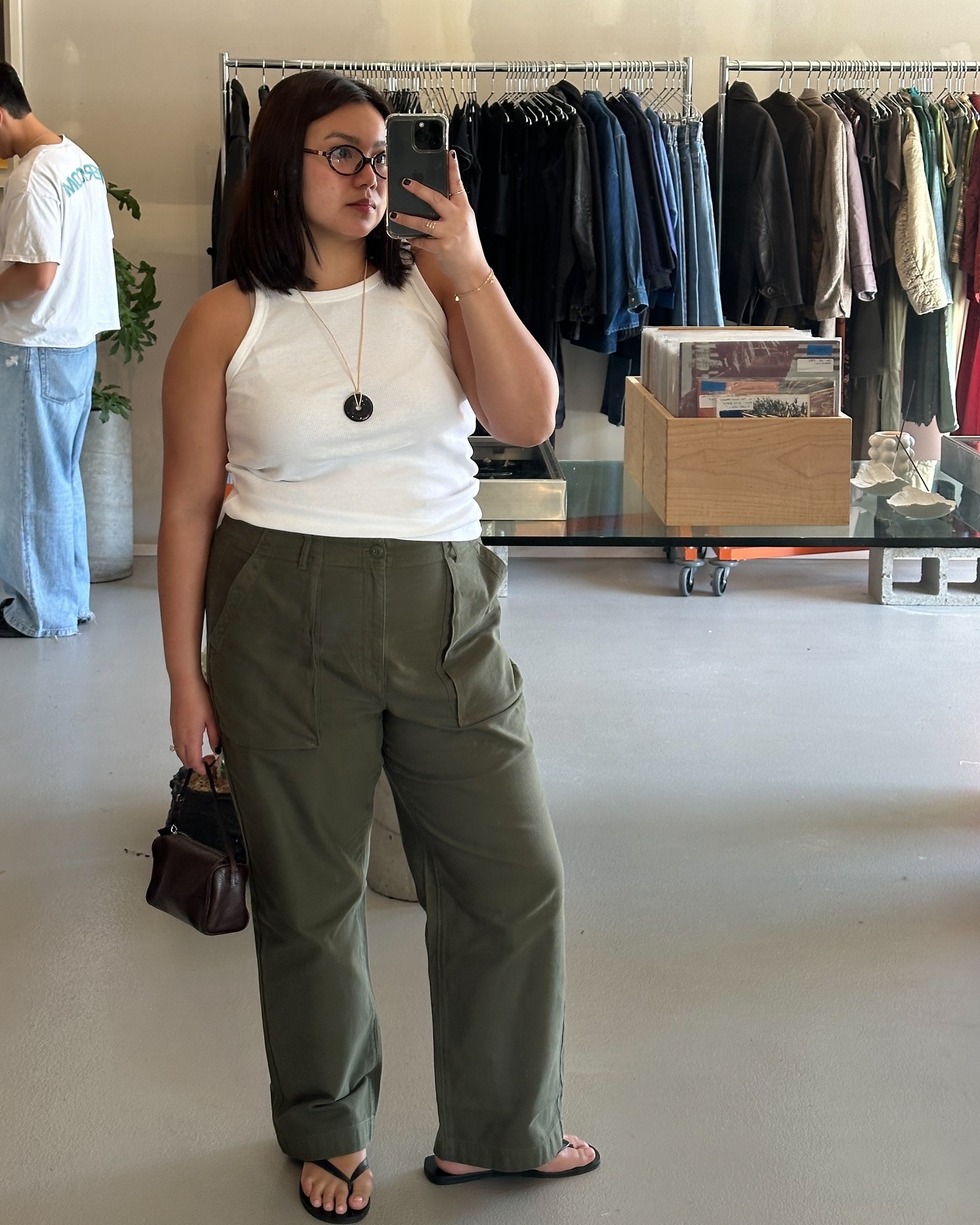 style influencer Marina Torres poses for a mirror selfie wearing oval optical eye glasses a white tank top, cord pendant necklace, relaxed green pants, and black flip-flop sandalss