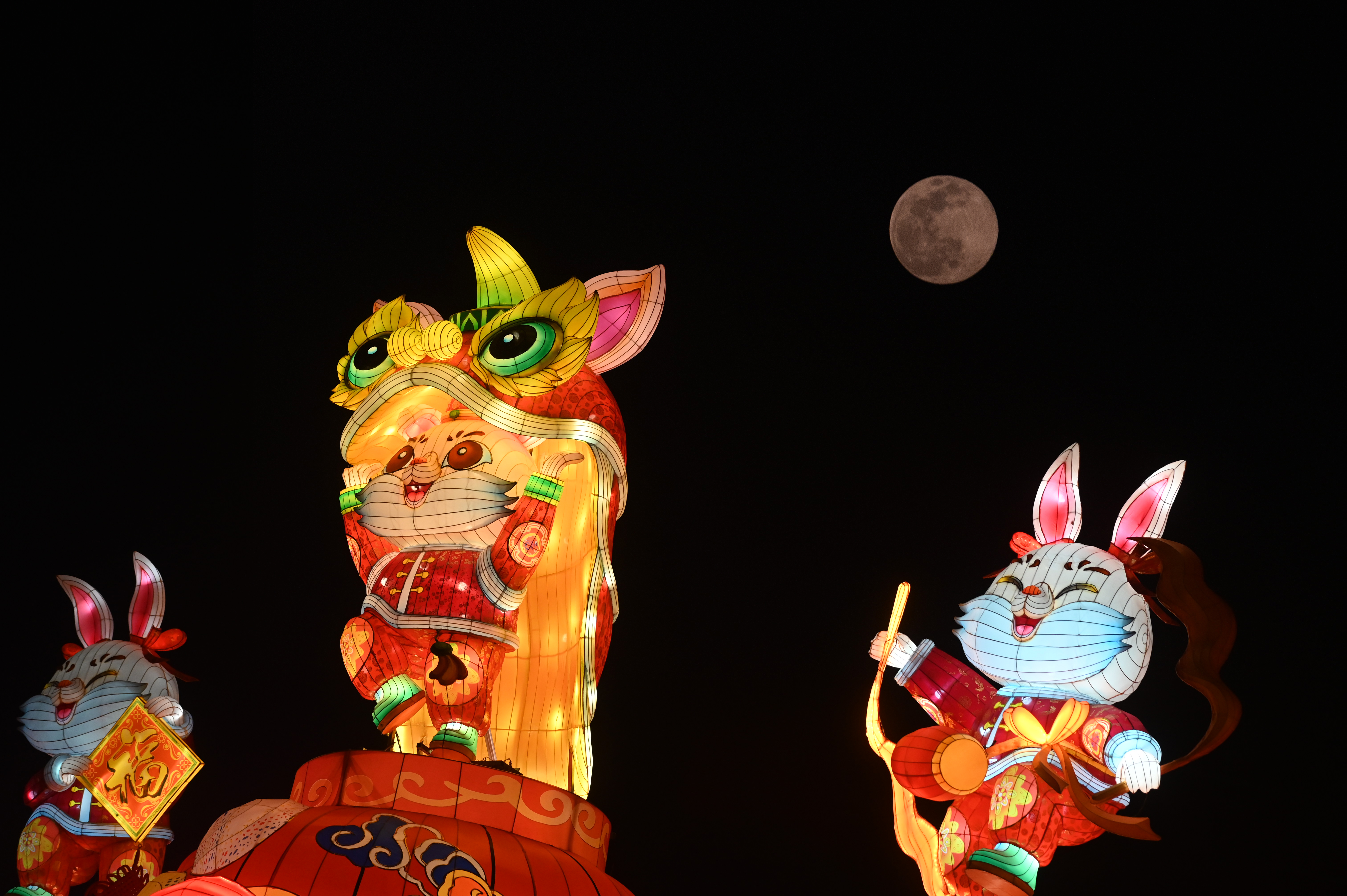 La luna llena se puede ver en el cielo nocturno detrás de tres linternas de colores con forma de animales sonrientes.