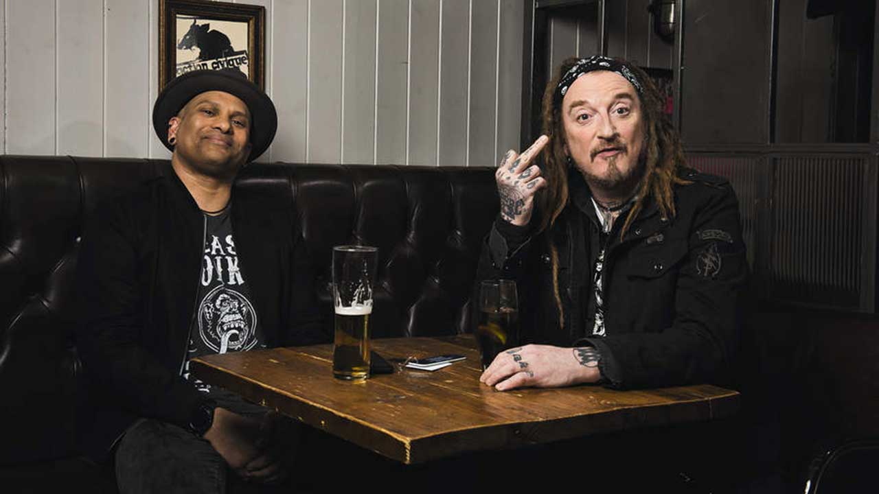 CJ Wildheart and Ginger Wildheart sitting in a pub, with Ginger extending a middle finger