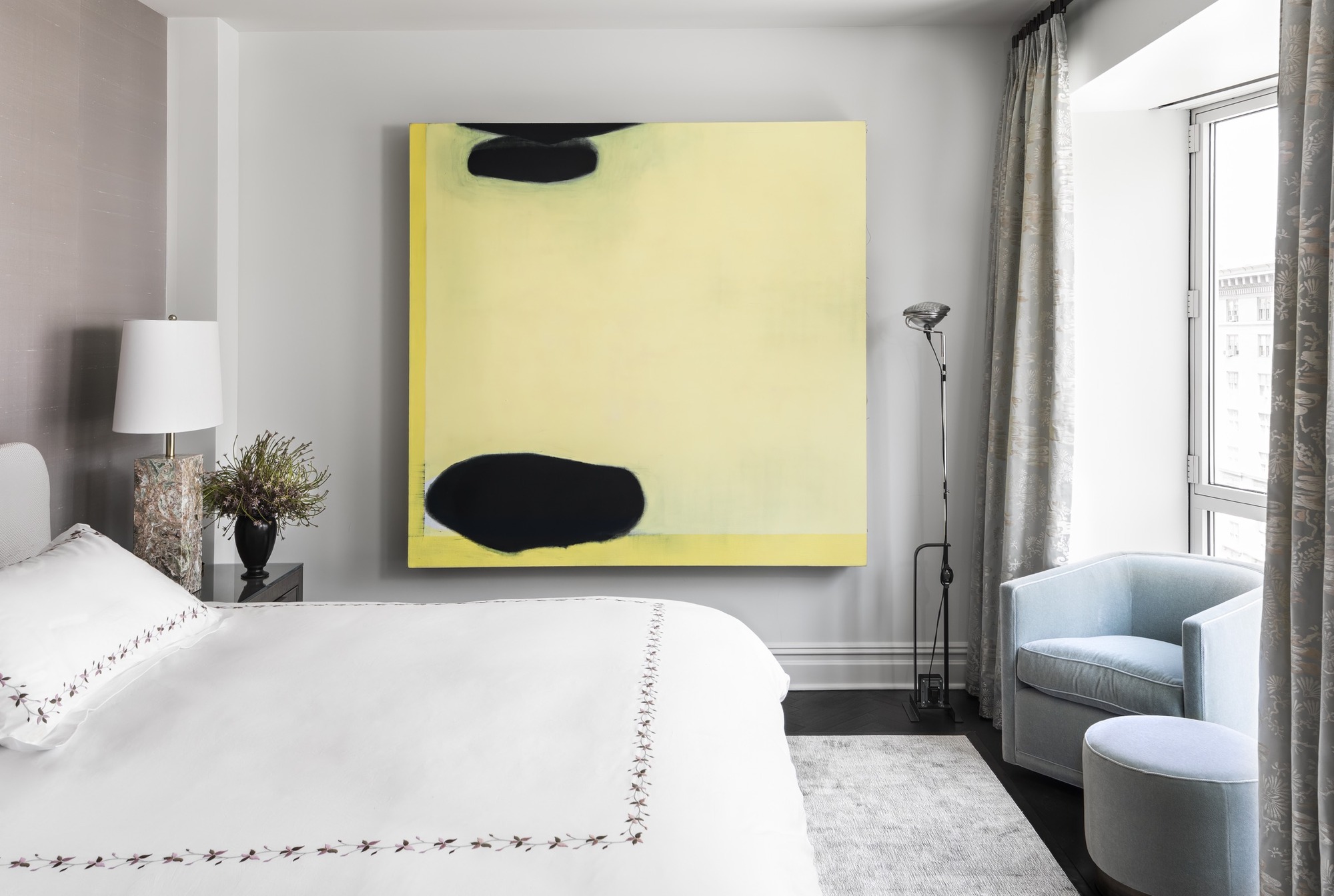 modern bedroom with large yellow artwork on wall, pink feature wall, white bedding with floral border, blue armchair and ottoman at foot of the bed in windowframe next to a floor lamp