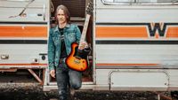 Myles Kennedy standing outside a trailer home with a guitar