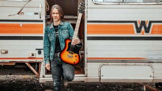 Myles Kennedy standing outside a trailer home with a guitar