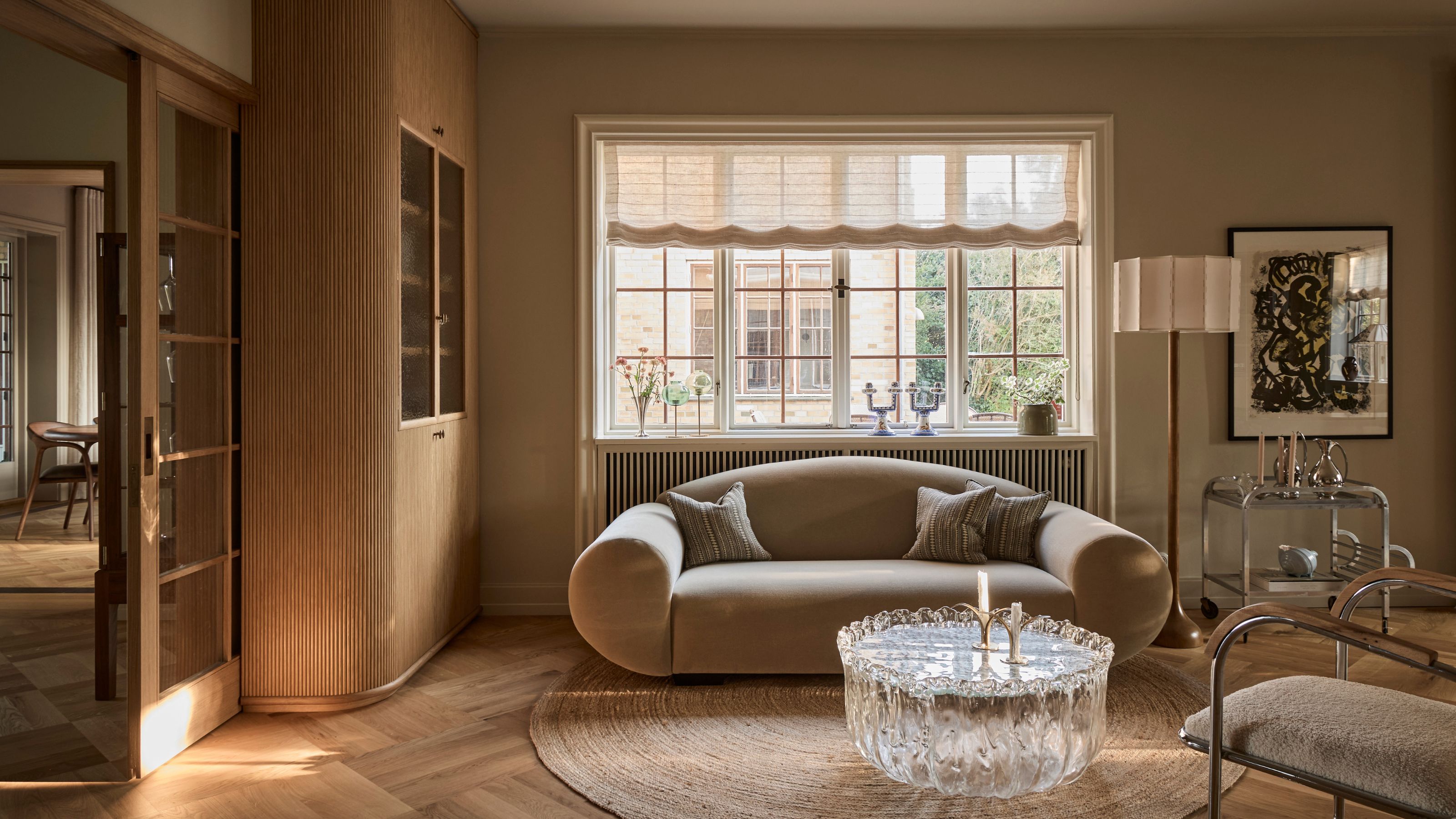 cozy living room with roman blinds, curved mohair sofa, round jute rug on light timber floor, glass coffee table, silver bar cart, floor lamp, artwork, white and chrome armchair and built-in timber bookcase, and sliding doors leading to the next room