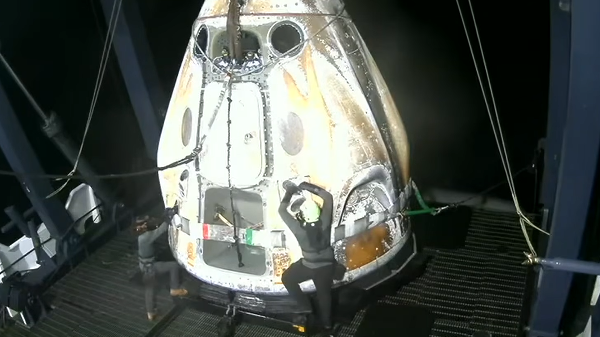 two people in hard hats climb onto a white cone-shaped capsule on the deck of a barge at sea