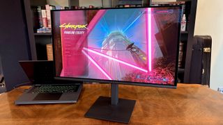 A widescreen monitor on a wooden desk displays the vibrant "Cyberpunk 2077: Phantom Liberty" menu, featuring neon lights and a futuristic cityscape.