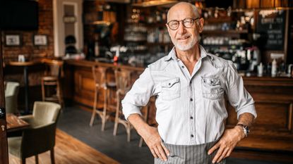 An older business owner stands with his hands on his hips, his coffee shop in the background.