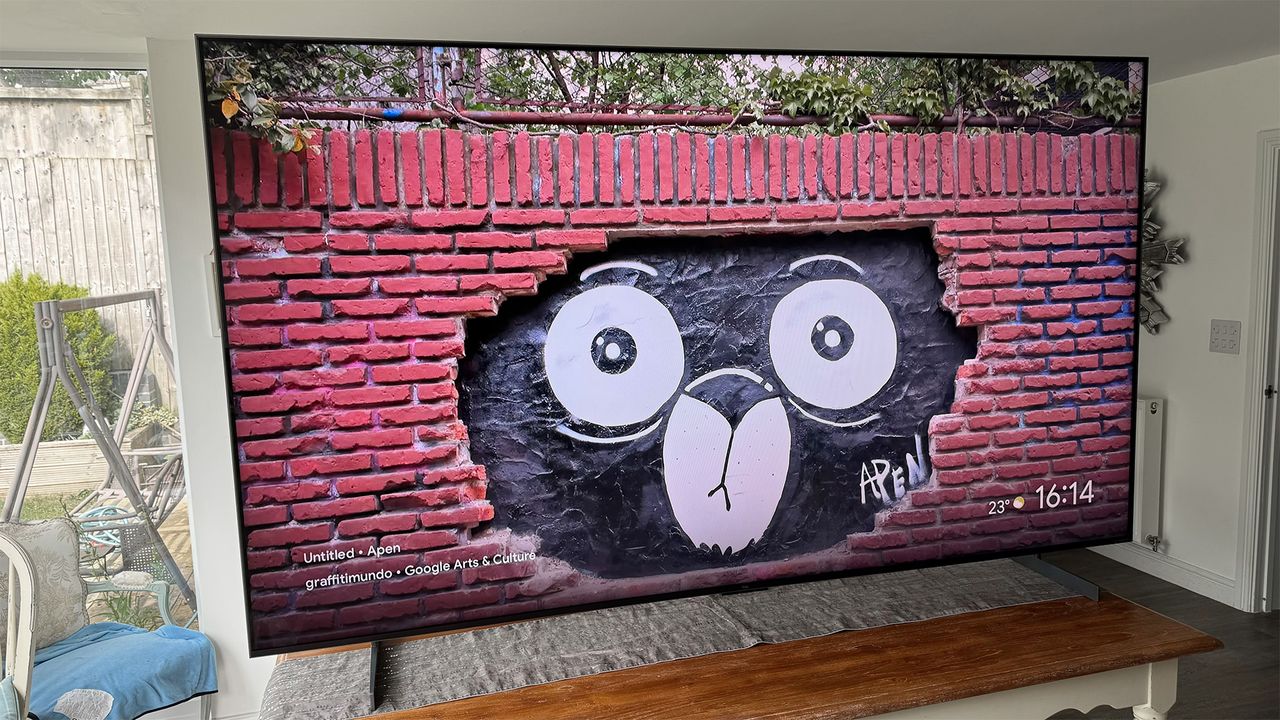 The TCL 98C7K 98-inch TV on a table in a living area, seen at a slight angle. On screen is a brick wall with an illustration of a cat's face in a section where the bricks are missing.