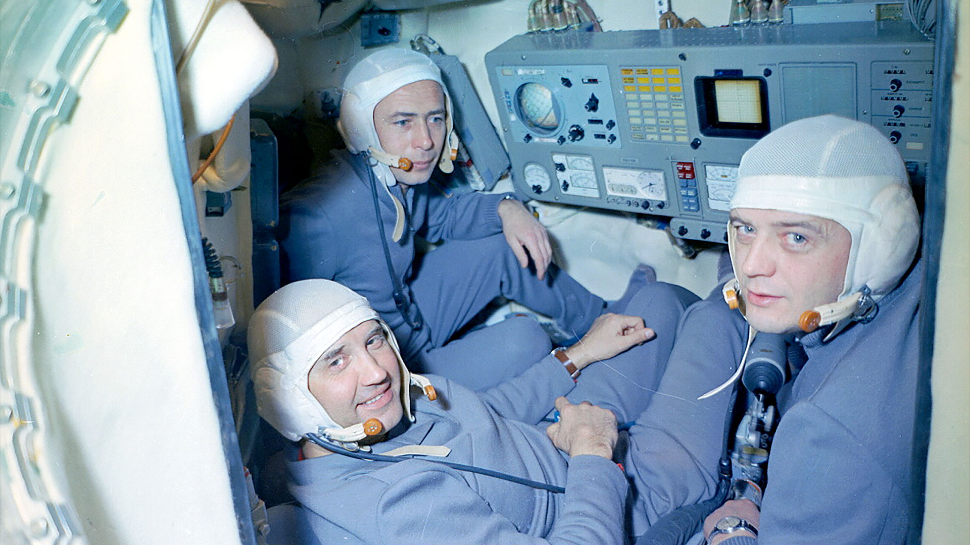 three men in blue coveralls and white caps sit inside a small space capsule mockup