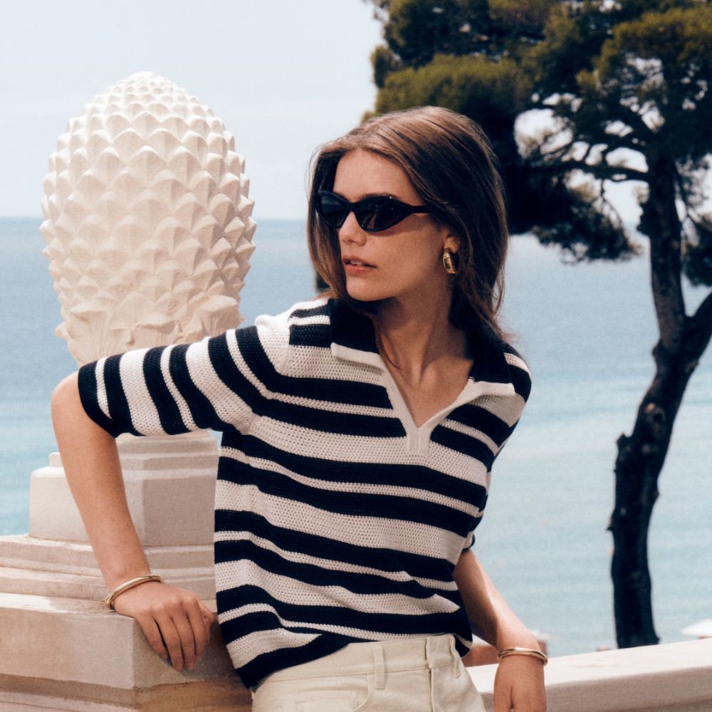 Young woman wearing monochrome outfit leaning on stone wall with sea and tree in background.