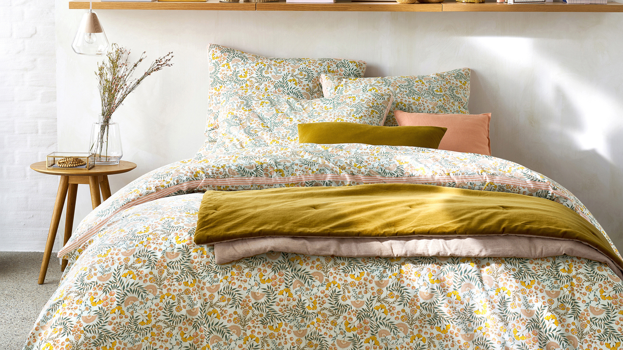 Floral bedding with a yellow throw on top. A cicular side table with dreid flowers and shelving unit above.