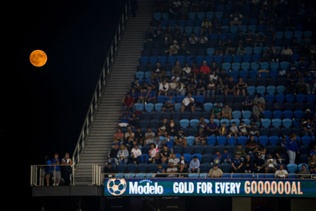 Uma lua laranja brilha no canto superior esquerdo da imagem, enquanto uma multidão de pessoas está sentada em bancos azuis à direita.