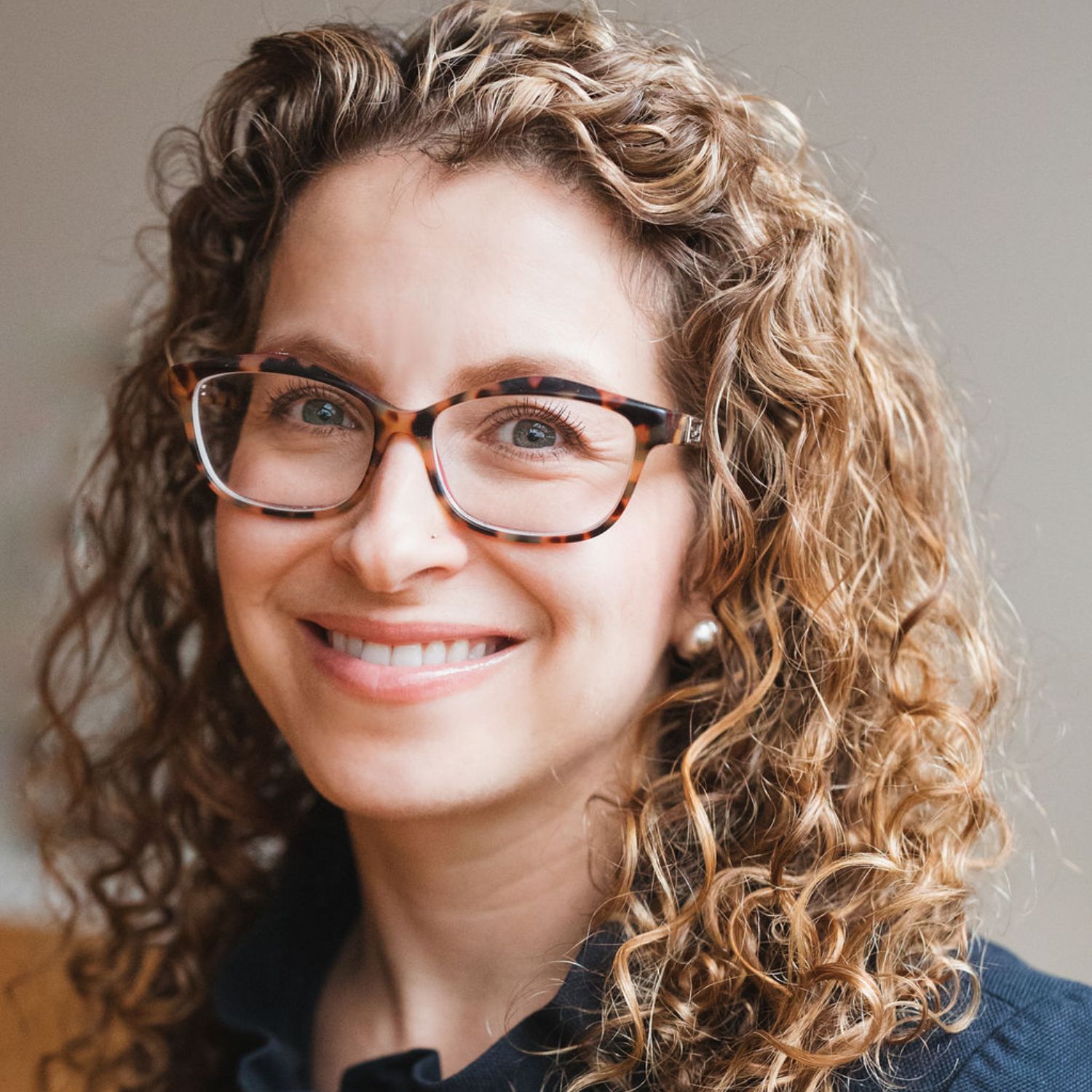 A headshot of a woman with curly brown hair, tortoise-shell glasses, and pearl earrings