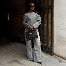 coco wears gray mono chrome outfit while holding a black handbag and wearing black pointed heels. 