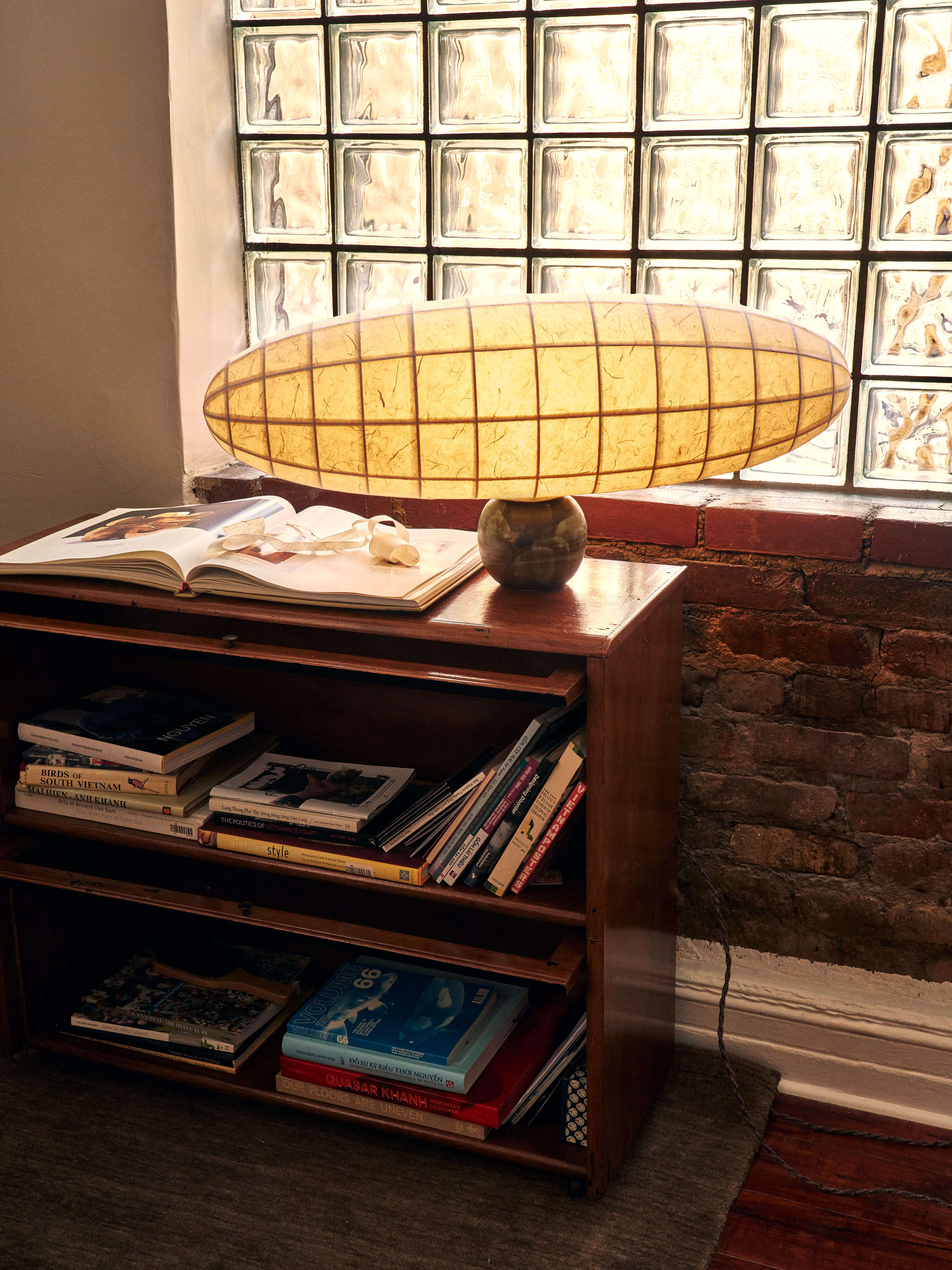 An oblong-shaped paper lantern with a checkered, horizontal surface resting on a globe-like stone sculpture, styled atop a wooden shelving unit in a naturally lit room with a glass block wall.