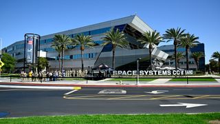 a crowd of people stand beside a blue rocket propellant tank outside an office building on a sunny day