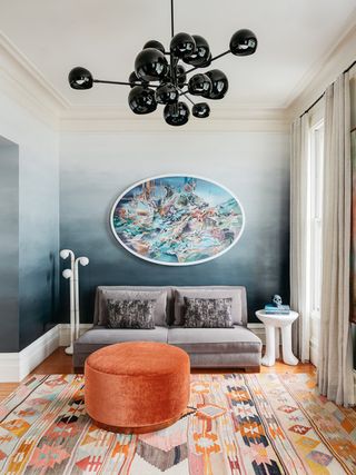 A guest bedroom with ombre gray/blue walls, a gray sleeper sofa, with an orange ottoman and rug