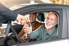 A salesperson hands car keys to a man sitting in the driver seat of a car.