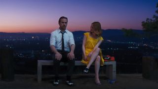 Ryan Gosling and Emma Stone sit on an overlook bench at dusk in Los Angeles, he's in a tie and she has a yellow dress on. 