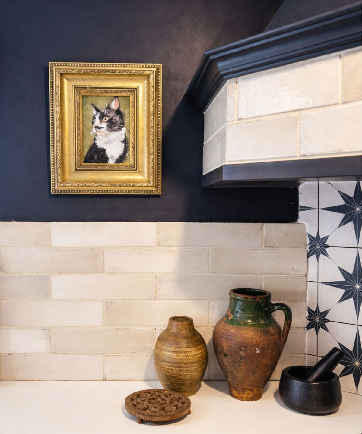 Navy kitchen corner with gold-framed cat portrait, cream tiles, white counter, rustic pottery, wooden trivet and black mortar and pestle.