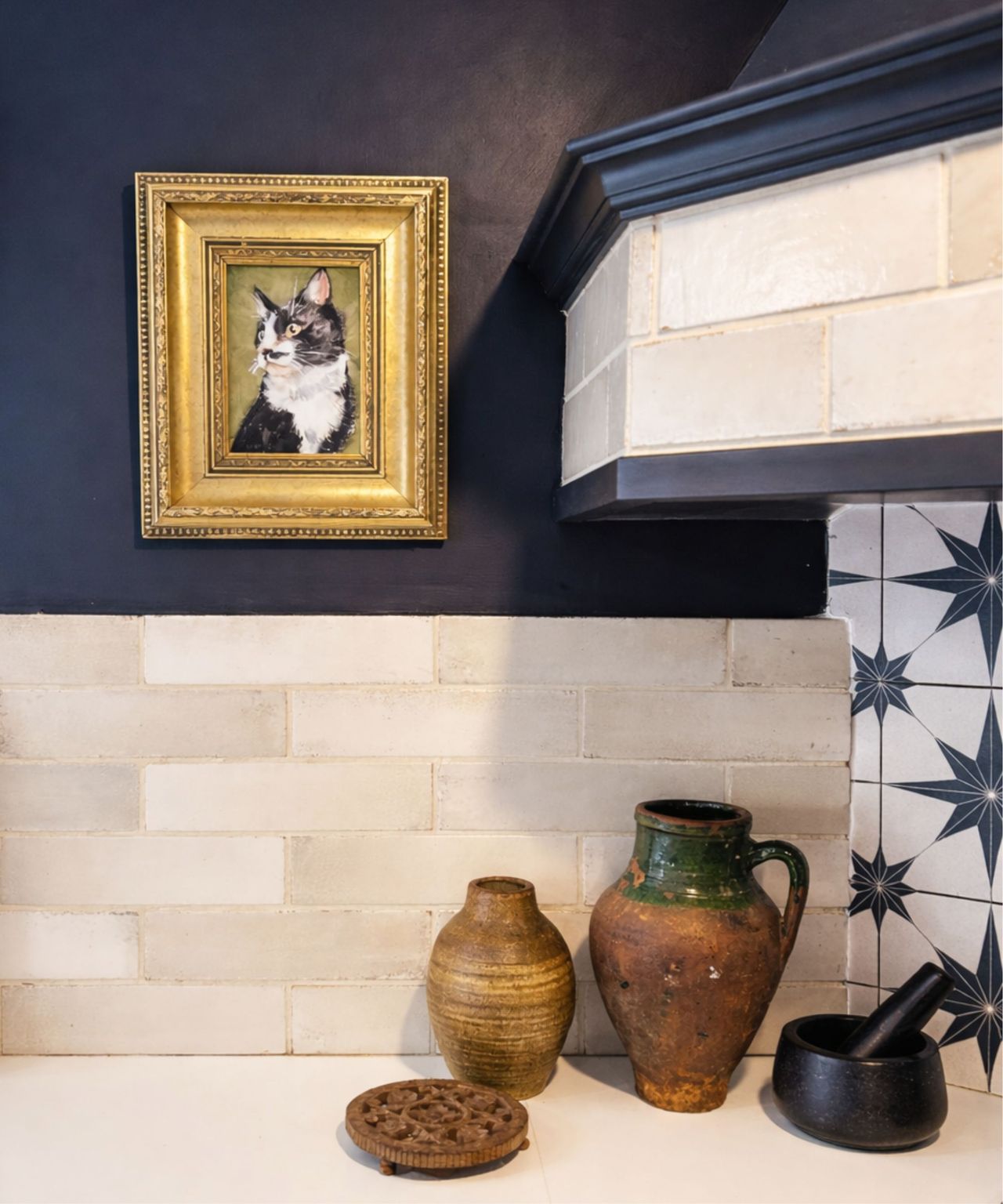 Navy kitchen corner with gold-framed cat portrait, cream tiles, white counter, rustic pottery, wooden trivet and black mortar and pestle.
