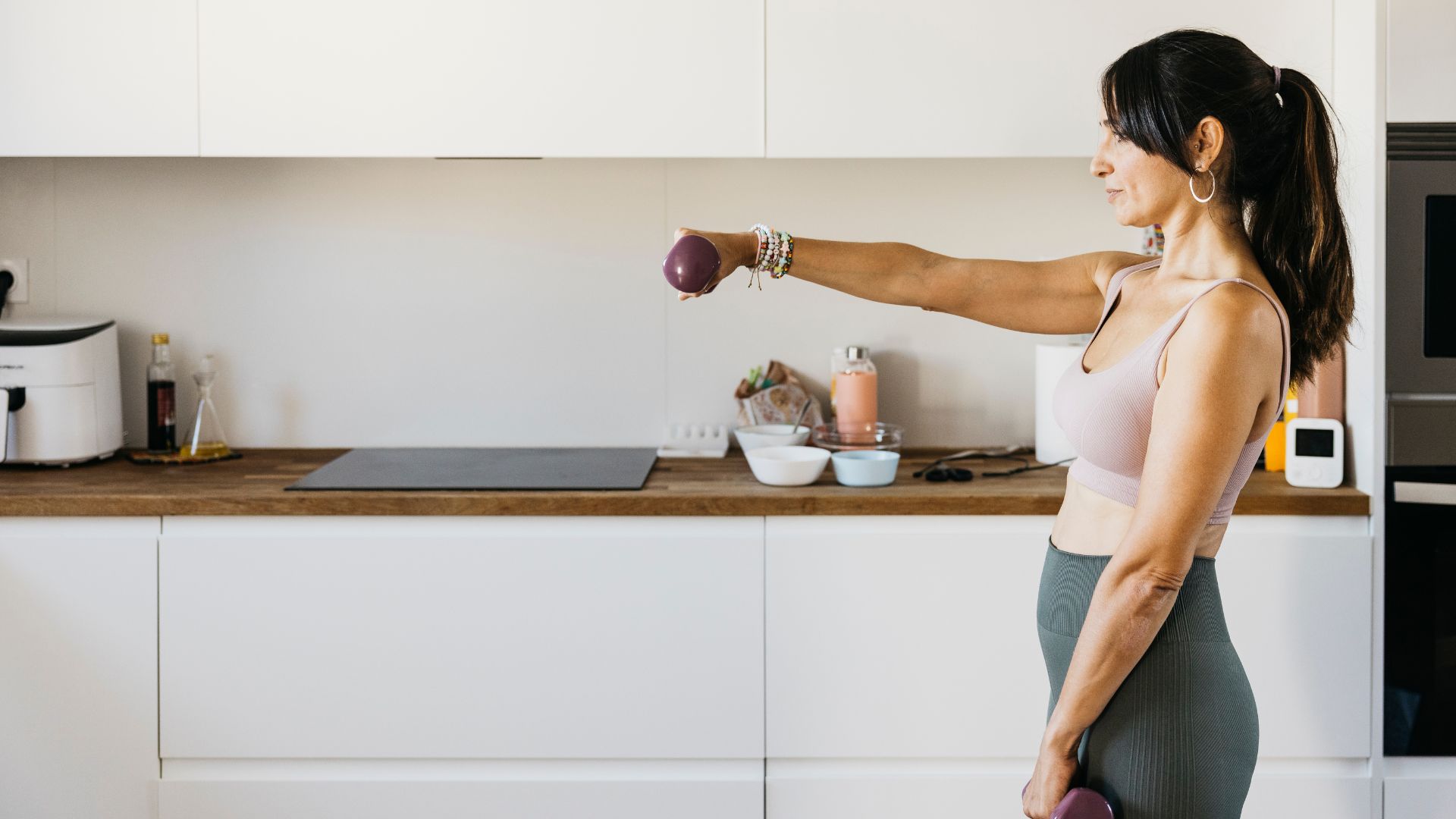 Woman doing lateral raises as part of a dumbbell workout at home in kitchen