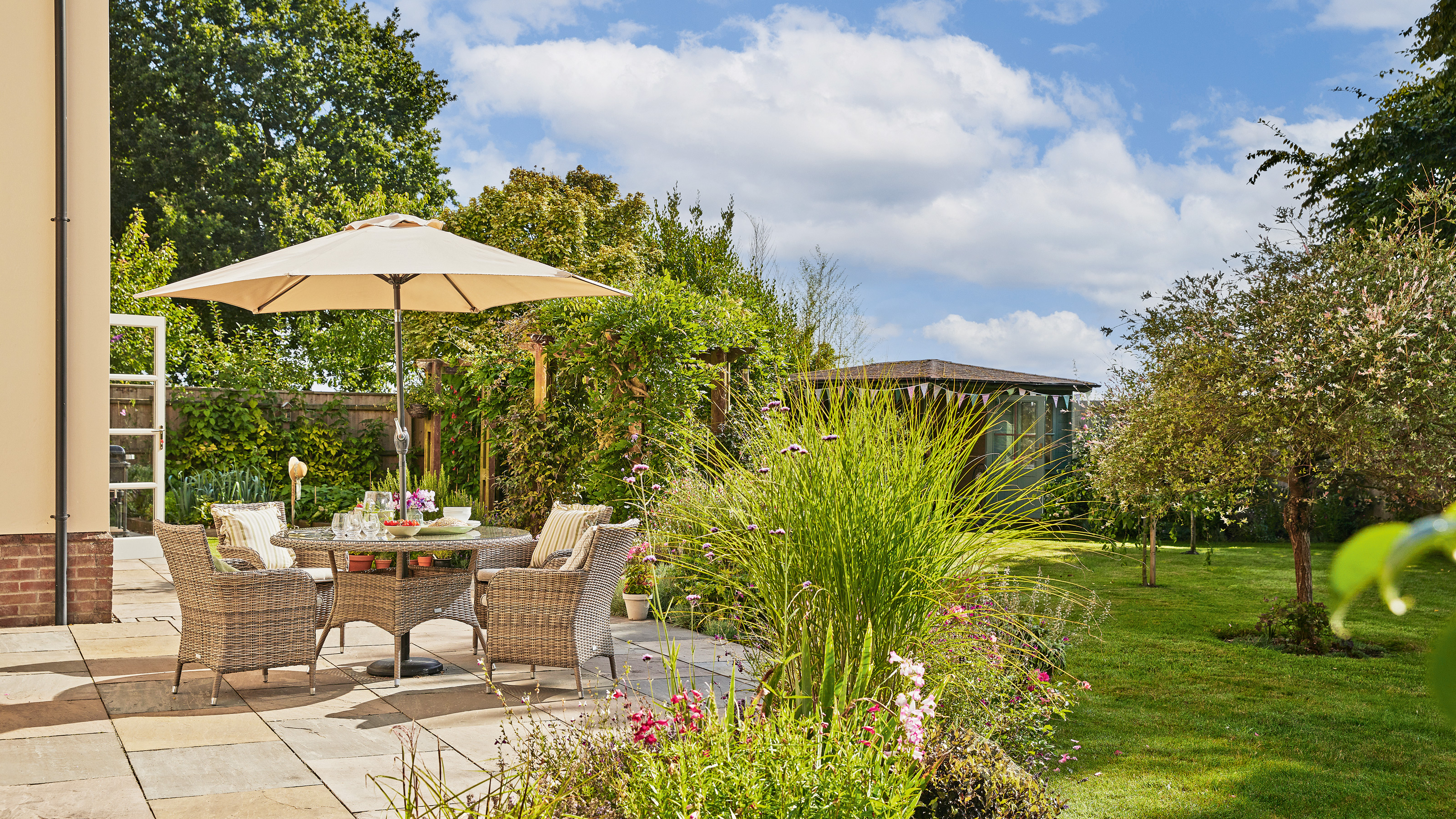 a patio with dining furniture and parasol alongside a lawn and summerhouse
