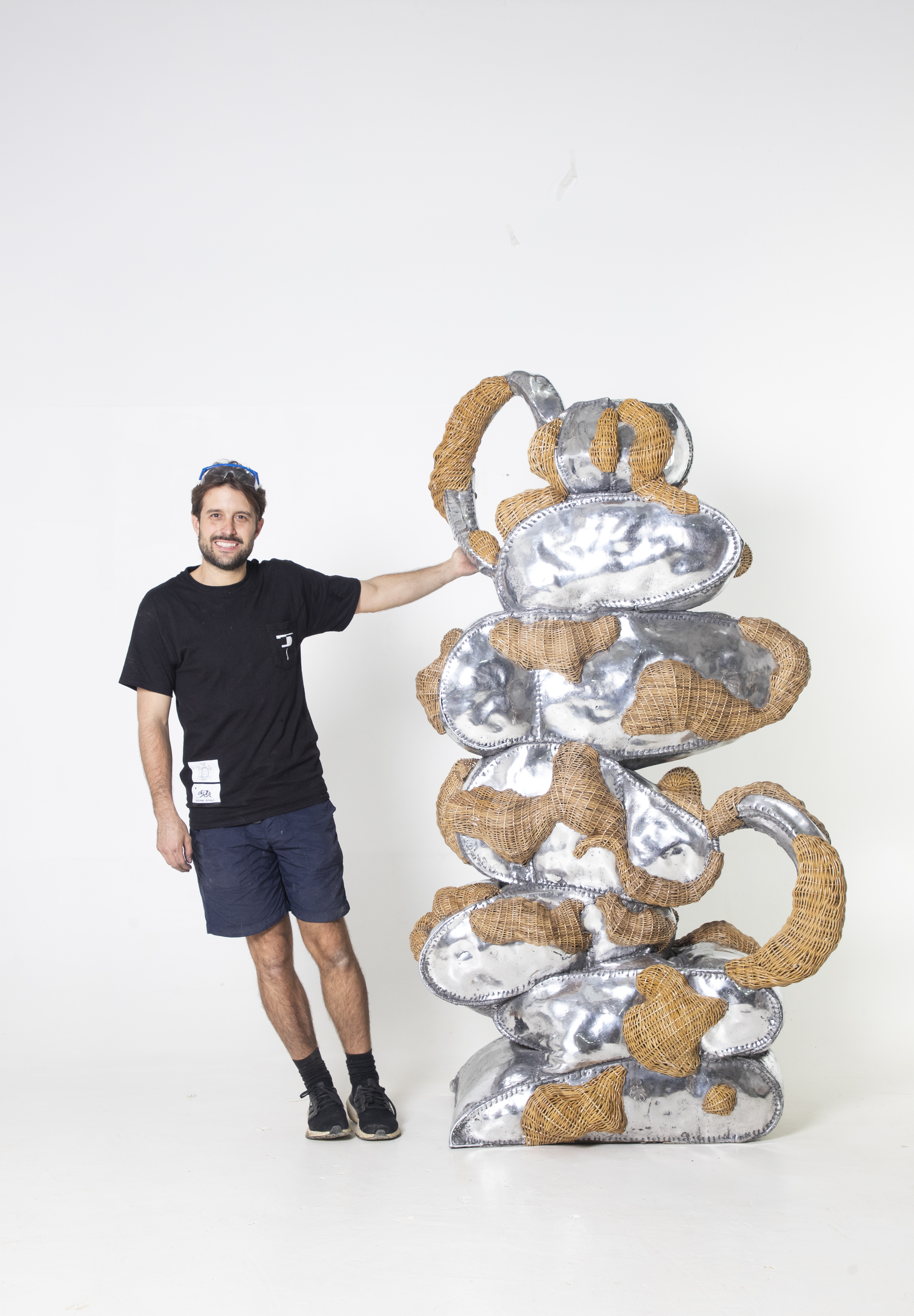 A man in a black T-shirt, navy blue shorts, and black trainers, with blue and white glasses over his short hair, stands next to a wicker and shiny silver sculpture while holding it with its left hand.