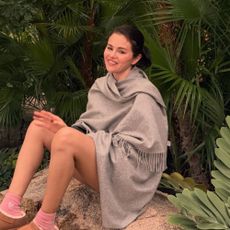 Selena Gomez sits on a large rock surrounded by trees and shrubbery wearing a scarf knit with Uggs and pink socks.