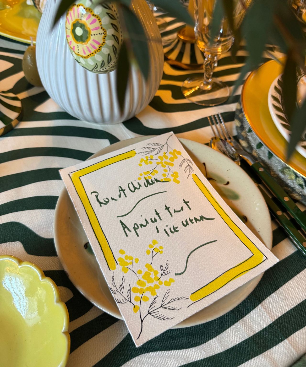 Menu card with yellow mimosa decoration on top of a plate on a table set for Easter with a green and yellow colour theme