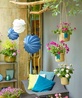 Garden pergola with bright pink geraniums in hanging baskets, blue and white hanging paper lanterns, and seating with bright blue and green cushions.