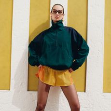 Model wearing yellow shorts and a dark green polo neck sweater and big sunglasses sold at Net-a-Porter in front of a yellow and white stripey wall
