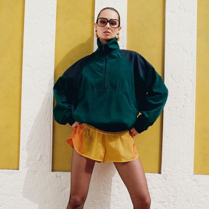 Model wearing yellow shorts and a dark green polo neck sweater and big sunglasses sold at Net-a-Porter in front of a yellow and white stripey wall