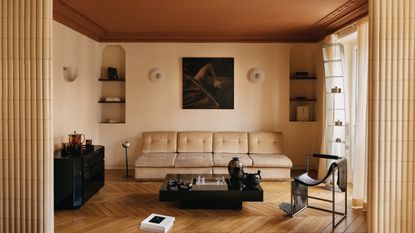 Image of a tan and beige living room with modern furniture and a large cream sofa. There are chrome accessories dotted throughout the room, and the ceiling is painted burnt orange.