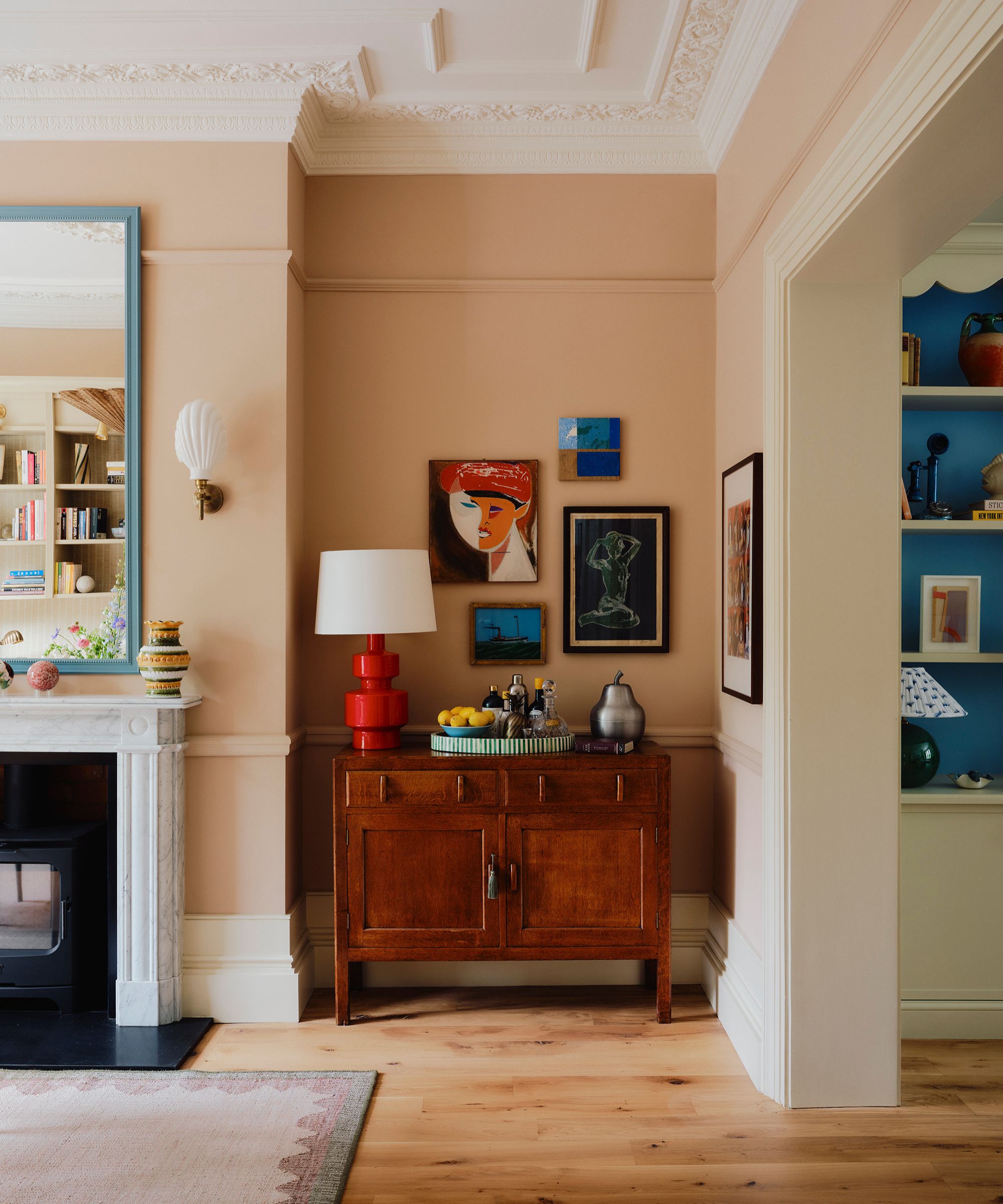 Living room with pale pink walls, wooden floors, and a marble fireplace with a large mirror above it, and an alcove with a dark wood vintage bar cabinet with a red lamp, barware, and artwork above it.
